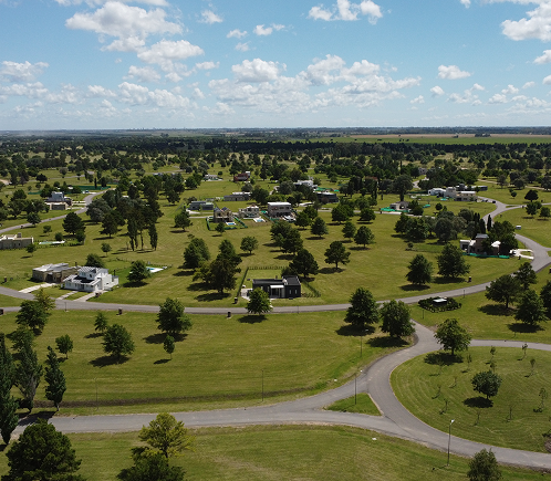 Vista aérea de un amplio barrio con caminos curvos, viviendas distribuidas entre grandes espacios verdes y árboles, bajo un cielo parcialmente nublado.