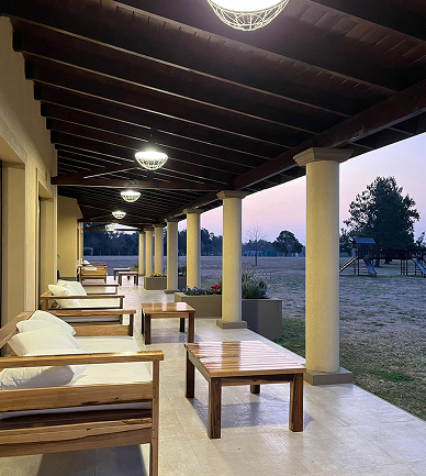 Galería techada con columnas, sillones de madera y mesas bajas, iluminada al atardecer, con vista a un amplio espacio verde exterior.