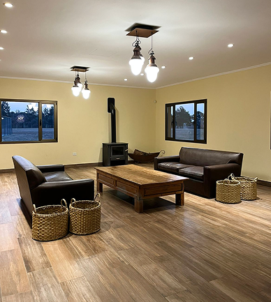 Sala de estar amplia con piso de madera, sillones marrones, mesa central rústica y estufa a leña con ventanas al exterior.
