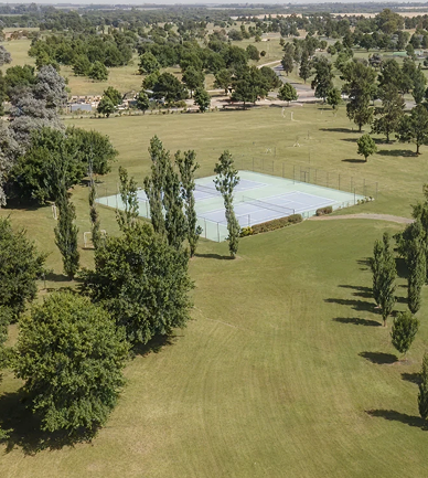 Vista aérea de cancha de tenis rodeada de amplios espacios verdes, árboles frondosos y paisaje campestre abierto.