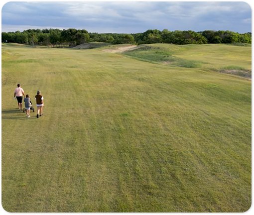 Amplio espacio verde con césped y árboles al fondo, donde tres personas caminan al aire libre bajo un cielo parcialmente nublado.