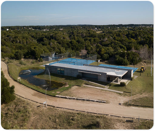 Vista aérea de canchas de tenis y un edificio deportivo rodeados de bosque, con edificios y el mar al fondo