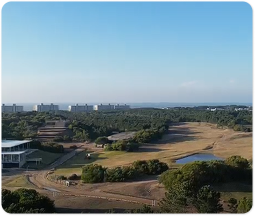 Vista aérea de canchas de tenis y un edificio deportivo rodeados de bosque, con edificios y el mar al fondo