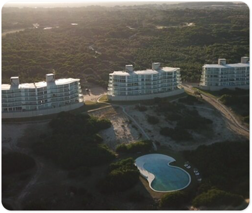 Vista aérea de canchas de tenis y un edificio deportivo rodeados de bosque, con edificios y el mar al fondo