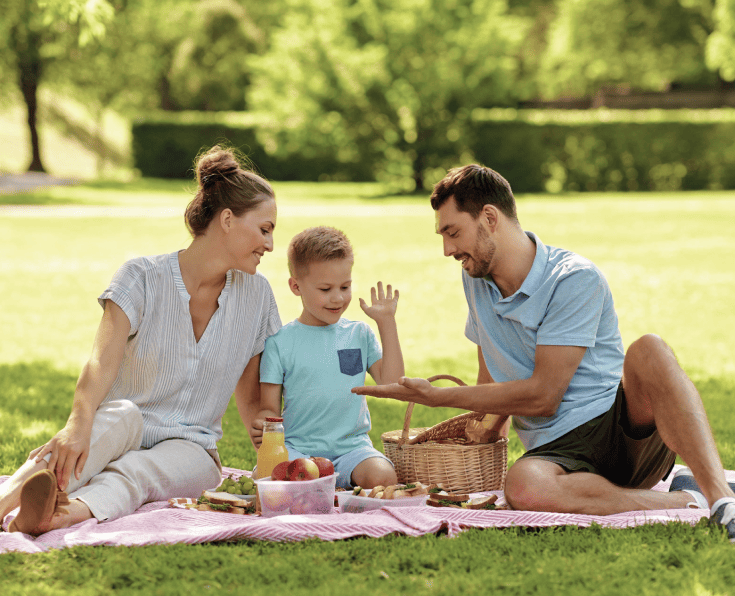 Familia disfrutando un picnic en el parque, sentados sobre una manta con comida y una canasta, mientras comparten un momento al aire libre rodeados de naturaleza.
